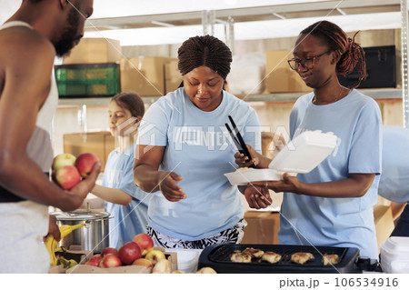 Friendly black women happily serving free food to the poor and needy african american man at a homeless shelter. Hungry, less fortunate male individual receives meal donation from hunger relief team. 106534916