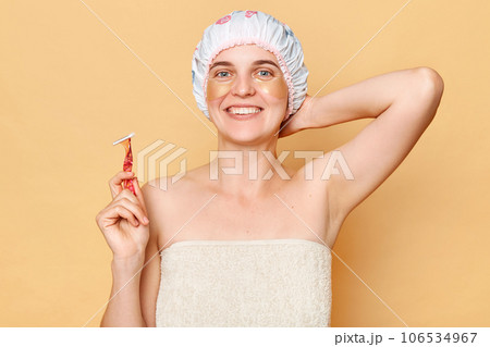 Attractive woman wearing shower cap standing isolated over beige background posing with patches under eyes shaving her armpit taking care of her skin. 106534967