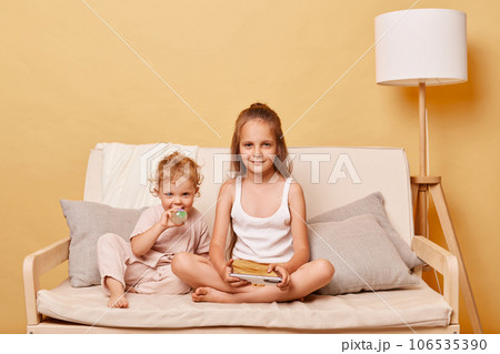 Relaxed charming little girls sisters using phone while sitting on sofa at home against beige wall looking at camera expressing positive emotions browsing internet together using smartphone. 106535390