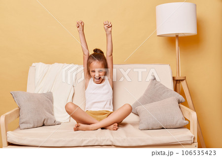 Ovedrjoyed happy positive optimistic little girl wearing casual clothing sitting on sofa at home against beige wall rejoicing her success raised clenched fists. 106535423