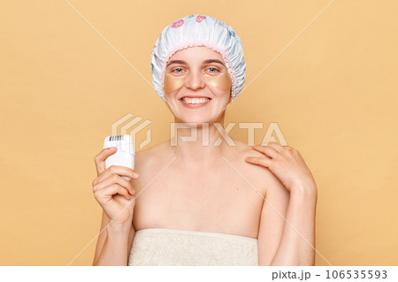 Optimistic woman wearing shower cap standing isolated over beige background holding depilator smiling happily posing in towel morning routine. 106535593