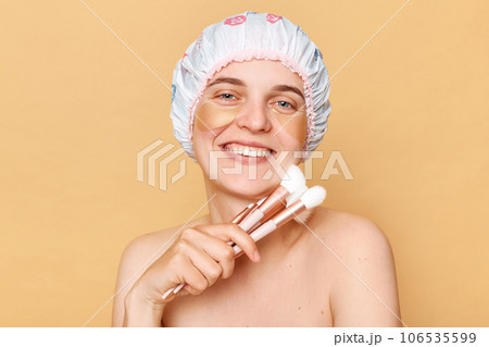 Beautiful attractive charming woman wearing shower cap standing isolated over beige background doing makeup in the morning after taking shower. 106535599