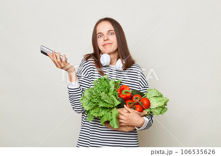 Fresh greenery and vegetables. Confused puzzled uncertain woman wearing striped casual shirt carrying vegetable and mobile phone using smartphone isolated over gray background Fresh greenery and vegetables. Confused puzzled uncertain woman wearing striped casual shirt carrying vegetable and mobile phone using smartphone isolated over gray background 106535626