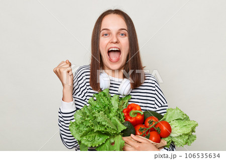 Extremely happy overjoyed woman wearing striped casual shirt carrying lettuce cabbage pepper avocado isolated over gray background clenched fist celebrating success. 106535634