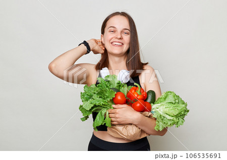 Delighted satisfied smiling woman wearing sportswear holding lettuce cabbage pepper avocado isolated over gray background buying organic food healthy nutrition. Delighted satisfied smiling woman wearing sportswear holding lettuce cabbage pepper avocado isolated over gray background buying organic food healthy nutrition. 106535691