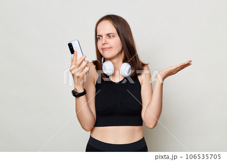 Puzzled confused slim woman with headphones wearing sportswear posing against gray background using mobile phone having problems with app. Puzzled confused slim woman with headphones wearing sportswear posing against gray background using mobile phone having problems with app. 106535705