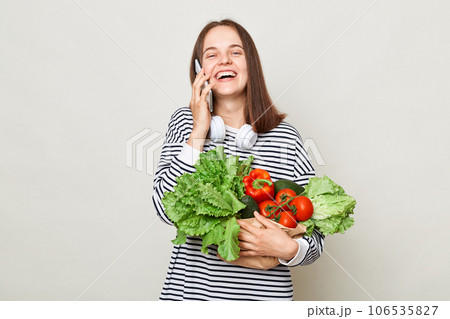 Happy laughing woman embraces fresh vegetables posing isolated over white background talking on mobile phone delivery of organic food. Happy laughing woman embraces fresh vegetables posing isolated over white background talking on mobile phone delivery of organic food. 106535827