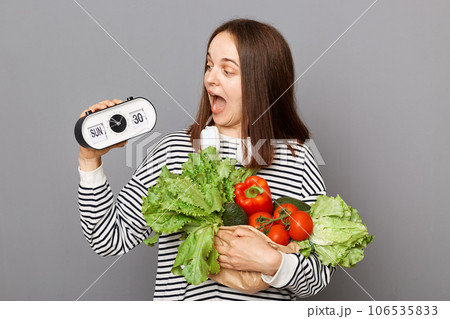 Time to healthy eating. Surprised woman embraces fresh vegetables posing isolated over gray background holding electronic clock looking at watch with amazed face. 106535833