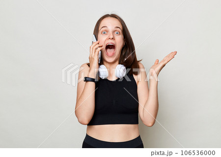 Shocked surprised woman wearing black top standing isolated over white background talking on cell phone hearing great news screaming with excitement. Shocked surprised woman wearing black top standing isolated over white background talking on cell phone hearing great news screaming with excitement. 106536000