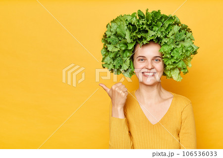 Fresh food care. Healthy eating habits. woman with lettuce hair style with green vegetables on her head isolated over yellow background pointing at advertisement area, copy space. Fresh food care. Healthy eating habits. woman with lettuce hair style with green vegetables on her head isolated over yellow background pointing at advertisement area, copy space. 106536033