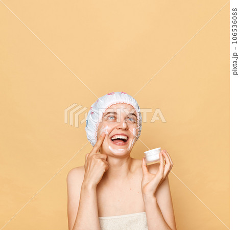 Laughing woman applies facial mask, puts nutritious cream, looking up at copyspace foe advertisement wearing shower cap and towel enjoying beauty treatment. 106536088