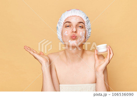Funny positive woman with pout lips, applies moisturising facial mask, holding nutritious cream in hands, wears shower cap sending air kisses looking at camera. Funny positive woman with pout lips, applies moisturising facial mask, holding nutritious cream in hands, wears shower cap sending air kisses looking at camera. 106536090