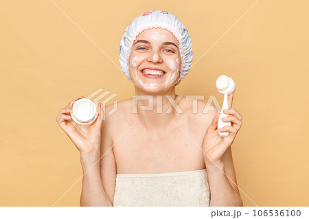 Attractive smiling happy pretty woman wearing shower cap taking bath doing beauty procedures holding cream and cleansing brush looking at camera with toothy smile. Attractive smiling happy pretty woman wearing shower cap taking bath doing beauty procedures holding cream and cleansing brush looking at camera with toothy smile. 106536100
