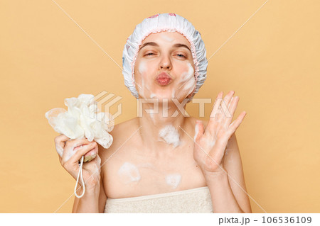 Relaxation self-care routine. Healthy hygiene habits. Positive optimistic young woman in shower cap and wrapped in towel sending air kissing isolated over beige background 106536109