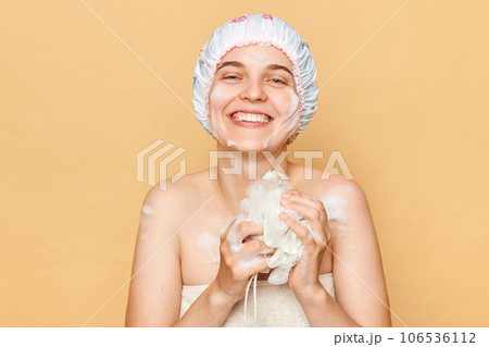 Satisfied positive smiling female dressed bath cap looking at camera with toothy smile holding sponge with foam taking shower in thew morning expressing happiness. Satisfied positive smiling female dressed bath cap looking at camera with toothy smile holding sponge with foam taking shower in thew morning expressing happiness. 106536112