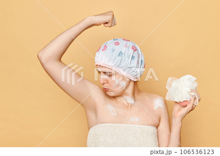 Young adult woman raised her arms smelling her armpit feeling unpleasant aromat frowning face being ready to take shower holding sponge wearing bath cap. Young adult woman raised her arms smelling her armpit feeling unpleasant aromat frowning face being ready to take shower holding sponge wearing bath cap. 106536123