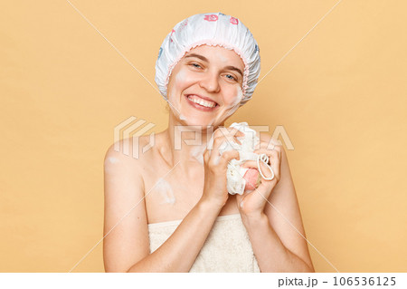 Pretty beautiful woman wearing shower cap standing isolated over beige background looking at camera holding sponge enjoying freshness after taking bath in the morning. 106536125
