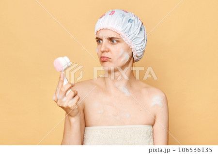 Confused puzzled woman wearing shower cap and wrapped in towel holding cleansing brush first time does't know how to use cosmetic tool standing isolated over beige background. 106536135