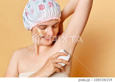 Smiling pretty woman using epilator isolated on beige background wearing bath cap showing her armpit smiling happily posing with patches under eyes. Smiling pretty woman using epilator isolated on beige background wearing bath cap showing her armpit smiling happily posing with patches under eyes. 106536154