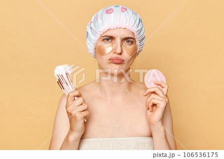 Sad upset girl holding makeup brushes and sponge looking at camera with unhappy face doing beauty procedures after morning shower standing isolated over beige background. 106536175