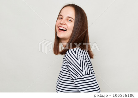 Attractive positive joyful woman with brown hair wearing striped shirt standing isolated over gray background looking at camera and laughing. Attractive positive joyful woman with brown hair wearing striped shirt standing isolated over gray background looking at camera and laughing. 106536186