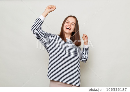 Extremely happy positive brown haired woman wearing striped shirt posing isolated over gray background standing with raised arms dancing celebrating holiday. Extremely happy positive brown haired woman wearing striped shirt posing isolated over gray background standing with raised arms dancing celebrating holiday. 106536187