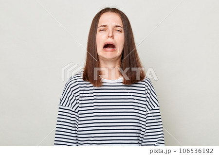 Unhappy sad brown haired woman wearing striped shirt posing isolated over gray background crying feeling despair hearing very bad news. Unhappy sad brown haired woman wearing striped shirt posing isolated over gray background crying feeling despair hearing very bad news. 106536192