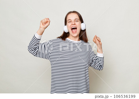 Positive happy cheerful brown haired woman wearing striped shirt posing isolated over gray background standing with headphones dancing with raised arms. Positive happy cheerful brown haired woman wearing striped shirt posing isolated over gray background standing with headphones dancing with raised arms. 106536199