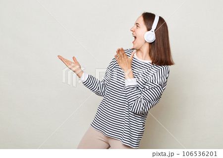 Positive woman with brown hair wearing striped shirt enjoying music on headphone showing advertisement area standing isolated over gray background. Positive woman with brown hair wearing striped shirt enjoying music on headphone showing advertisement area standing isolated over gray background. 106536201