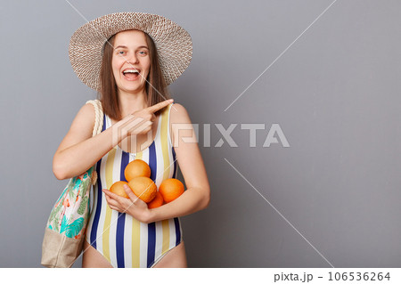 Smiling delighted woman wearing striped colorful swimwear standing with bag and oranges isolated over gray background pointing aside at copy space for promotional text. Smiling delighted woman wearing striped colorful swimwear standing with bag and oranges isolated over gray background pointing aside at copy space for promotional text. 106536264