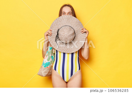 Funny woman wearing swimsuit isolated on yellow background covering half of her face with her straw hat looking with satisfied expression relaxing at exotic tour. Funny woman wearing swimsuit isolated on yellow background covering half of her face with her straw hat looking with satisfied expression relaxing at exotic tour. 106536285