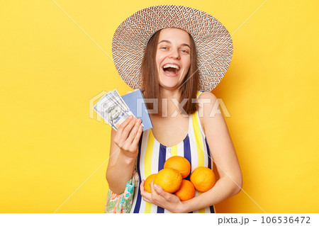 Extremely happy cheerful woman wearing striped swimsuit and straw hat isolated yellow background holding oranges passport and dollar banknotes fro traveling. 106536472