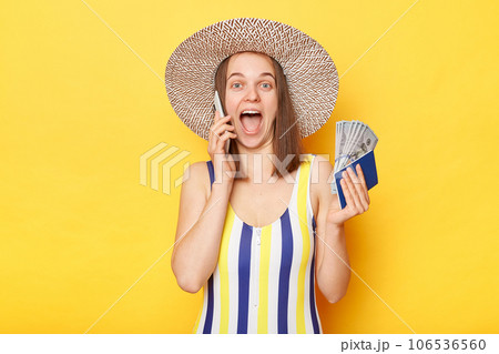 Unexpected vacation. Happy amazed woman on resort dressed in striped swimwear and straw hat posing isolated over yellow background holding passport and money talking phone boasting her vacation. Unexpected vacation. Happy amazed woman on resort dressed in striped swimwear and straw hat posing isolated over yellow background holding passport and money talking phone boasting her vacation. 106536560