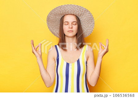 Calm woman on resort dressed in striped swimwear and straw hat posing isolated over yellow background practicing yoga meditating on resort keeps eyes closed. 106536568