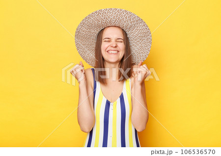 Cheerful overjoyed woman wearing striped swimming suit and sun hat isolated on yellow background standing with clenched fists rejoicing her tropical vacation. 106536570