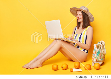 Hard working attractive woman wearing one piece swimming suit posing isolated over yellow background working on resort sitting with laptop enjoying her remote work. Hard working attractive woman wearing one piece swimming suit posing isolated over yellow background working on resort sitting with laptop enjoying her remote work. 106536611