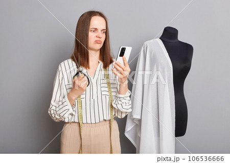 Sad upset professional female dressmaker expressing sorrow poising isolated over gray background using mobile phone checking orders in internet, has no work. 106536666