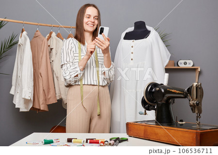 Portrait of beautiful woman seamstress holding phone in hand, tailor typing message on smartphone, searching patterns in internet, dressmaker working in workshop. Portrait of beautiful woman seamstress holding phone in hand, tailor typing message on smartphone, searching patterns in internet, dressmaker working in workshop. 106536711