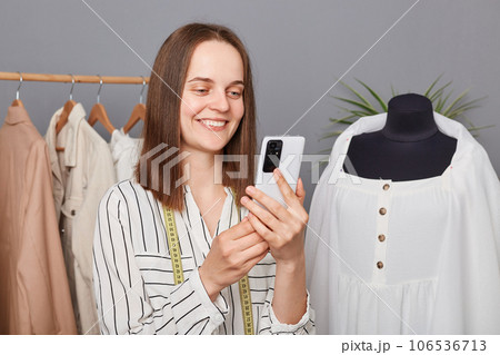 Smiling attractive young professional dressmaker using smartphone while working in atelier, searching new ideas in internet, browsing networks, fashion blog, sewing dresses for new collection. Smiling attractive young professional dressmaker using smartphone while working in atelier, searching new ideas in internet, browsing networks, fashion blog, sewing dresses for new collection. 106536713