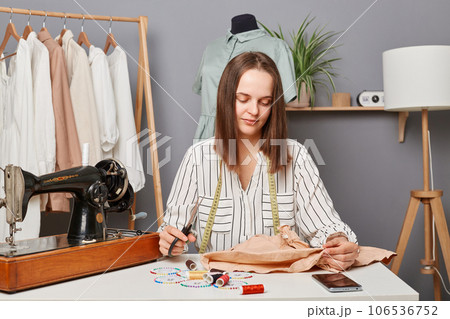 Young focused concentrated woman sewer sitting at her workplace in atelier near sewing machine with scissors in hands, thinking how to alter an old dress or shirt. Young focused concentrated woman sewer sitting at her workplace in atelier near sewing machine with scissors in hands, thinking how to alter an old dress or shirt. 106536752