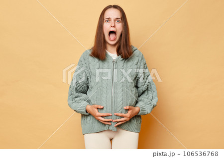 Shocked scared pregnant woman wearing knitted warm sweater standing isolated over beige background touching her belly screaming with fear feeling contractions. 106536768