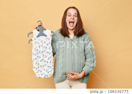 Excited amazed pregnant woman wearing knitted warm sweater holding kid's bodysuits standing isolated over beige background showing cotton clothes for her baby for low price. 106536775