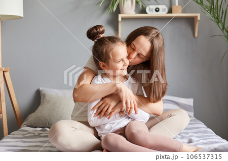 Charming little child girl congratulating mom with mother's day, daughter sitting in bed with mommy, gentle moment in morning, woman kissing her kid with love. 106537315