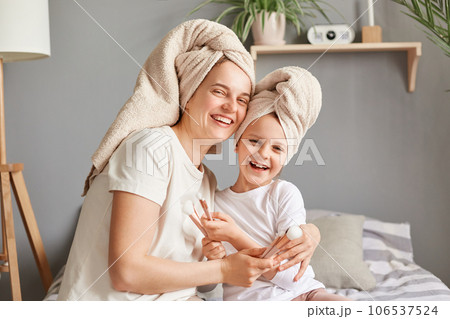 Joyful woman hugging her little kid mother and daughter in towels posing in bedroom looking at camera expressing happiness beauty procedures together. Joyful woman hugging her little kid mother and daughter in towels posing in bedroom looking at camera expressing happiness beauty procedures together. 106537524