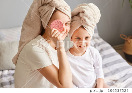 Happy child with mother sitting on bed in towels after morning shower having fun woman covering eye with cosmetic sponge family doing beauty procedures. 106537525
