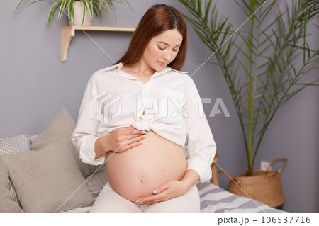 Beautiful expectant mother sitting on bed, touching her bare tummy with affection, happily waiting for her baby's arrival, wearing white shirt looking at her belly. Beautiful expectant mother sitting on bed, touching her bare tummy with affection, happily waiting for her baby's arrival, wearing white shirt looking at her belly. 106537716