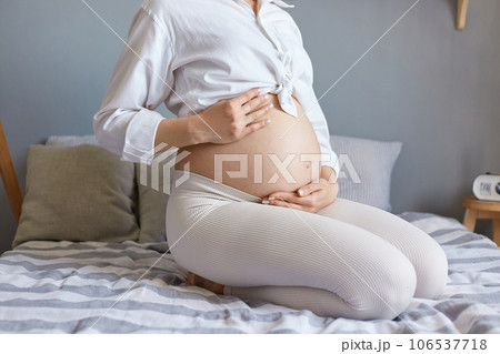 Healthy pregnancy. Expecting childbirth. Unrecognizable unknown pregnant woman wearing white clothing sitting in bed in light bedroom holding her bare belly. Healthy pregnancy. Expecting childbirth. Unrecognizable unknown pregnant woman wearing white clothing sitting in bed in light bedroom holding her bare belly. 106537718