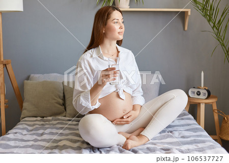Attractive dreamy pregnant woman with bare belly wearing white clothing sitting in bed in light bedroom, touching her belly and caring about her health, drinking fresh water. 106537727