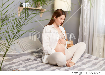 Pregnant woman sitting comfortably in her bedroom, young caucasian mother-to-be with bare belly enjoying quiet moment, anticipating the birth of her baby. Pregnant woman sitting comfortably in her bedroom, young caucasian mother-to-be with bare belly enjoying quiet moment, anticipating the birth of her baby. 106537748