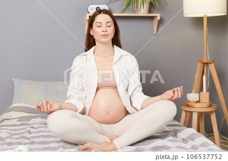 Calm beautiful pregnant brown haired caucasian woman expecting baby sitting in bed sitting with closed eyes meditating practicing yoga. Calm beautiful pregnant brown haired caucasian woman expecting baby sitting in bed sitting with closed eyes meditating practicing yoga. 106537752